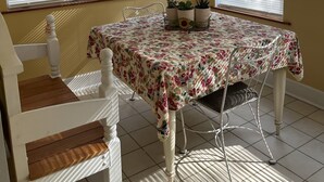 Un coin repas avec une table recouverte d’une nappe à motifs floraux, des chaises blanches et une petite plante servant de centre de table.