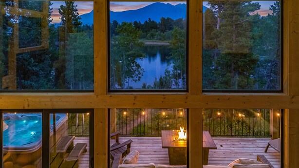 A cabin with multiple windows overlooking a lake and forest, a hot tub, and a deck with outdoor seating and a fire pit. - Lakefront - Alpine Views - Daily Moose (Black Hawk)