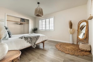 Ein Schlafzimmer mit einem Bett, einer Hängelampe, einem Fenster mit Jalousien, einem Spiegel und einer Zimmerpflanze.