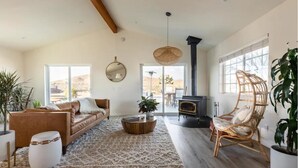 A spacious living room with a sofa, coffee table, and a fireplace. There are large windows and a view of the outdoors. - The Blue Cactus Ranch - Pool and Spa (Joshua Tree)