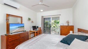A bedroom with a bed, a desk with a computer, a TV, a dresser, and a balcony with a view of greenery. - Modern 2 bed townhouse 200 metre from beach - your home away from home (Port Douglas)