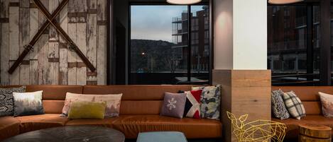 A modern lounge area with a leather sofa, a round table, and a view of buildings through large windows.