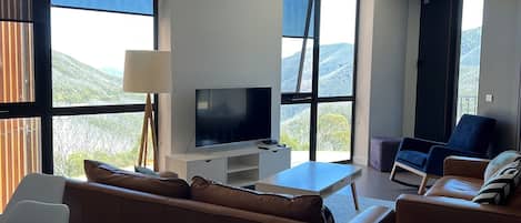 A modern living room with a flat-screen TV, comfortable seating, and large windows offering a view of the outdoors.