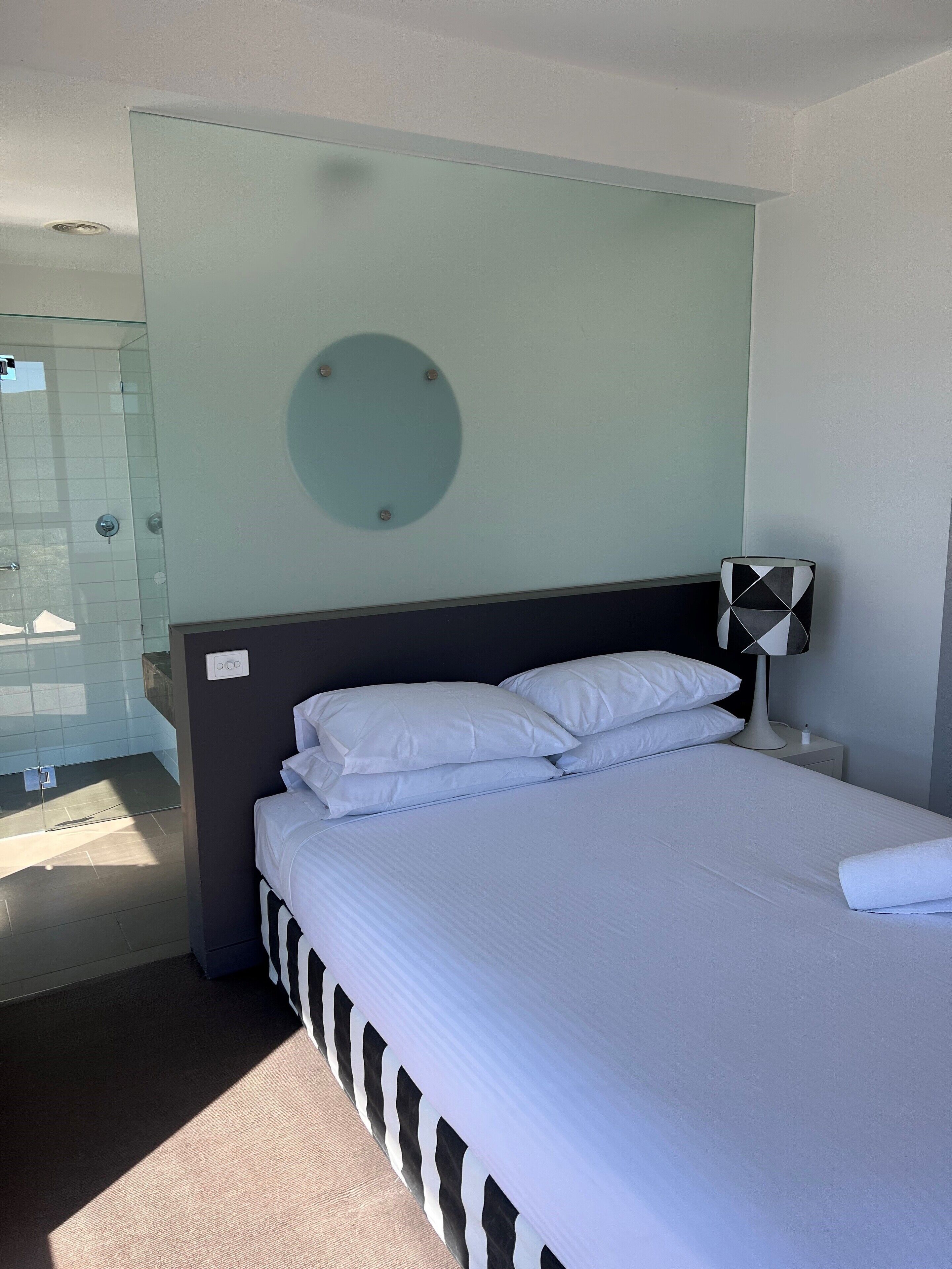 A hotel room with a bed, a nightstand, a lamp, and a bathroom visible in the background.