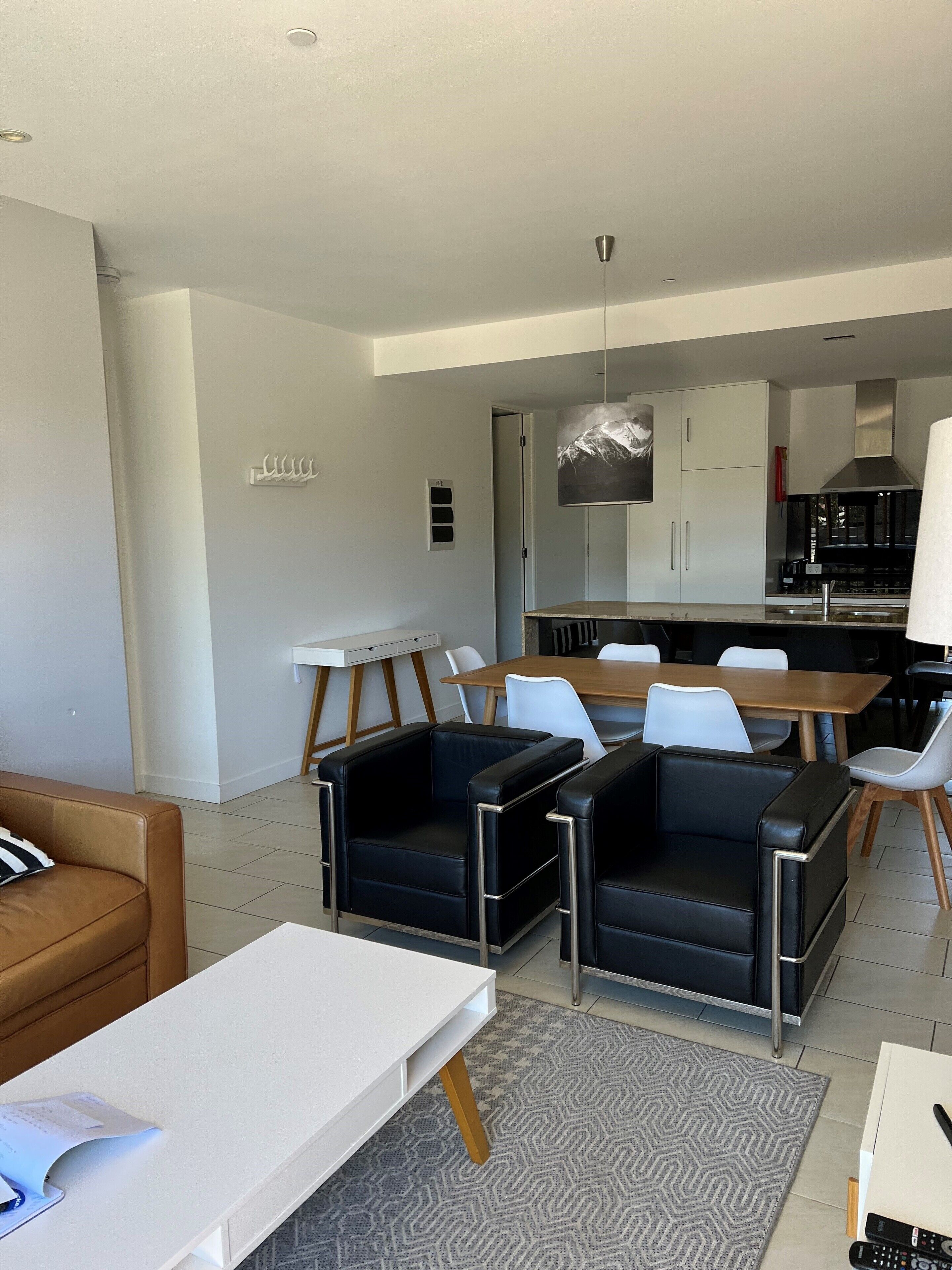 A modern living room with a brown leather sofa, a white coffee table, and black leather armchairs. There is a dining area with a wooden table and white chairs. The kitchen is visible in the background with white cabinets and a black stove.