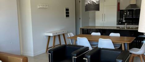 A modern living room with a brown leather sofa, a white coffee table, and black leather armchairs. There is a dining area with a wooden table and white chairs. The kitchen is visible in the background with white cabinets and a black stove.