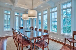 A spacious dining room with a large wooden table, multiple chairs, and large windows offering a view of the outdoors.