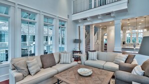 A spacious living room with a high ceiling, large windows, a built-in staircase, and a view of the outside balcony.