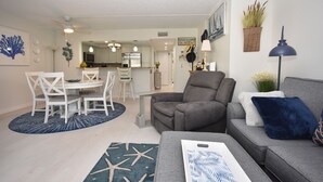 Un salon moderne avec un canapé gris, une table à manger entourée de chaises blanches et une cuisine en arrière-plan.
