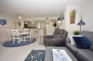 A modern living room with a grey sofa, a dining table with white chairs, and a kitchen in the background.