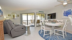 Un salon avec un fauteuil inclinable gris, une table à manger blanche avec des chaises, un ventilateur de plafond et un téléviseur fixé au mur.