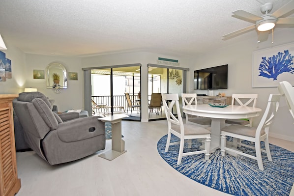 A living room with a gray recliner, a white dining table with chairs, a ceiling fan, and a TV mounted on the wall.