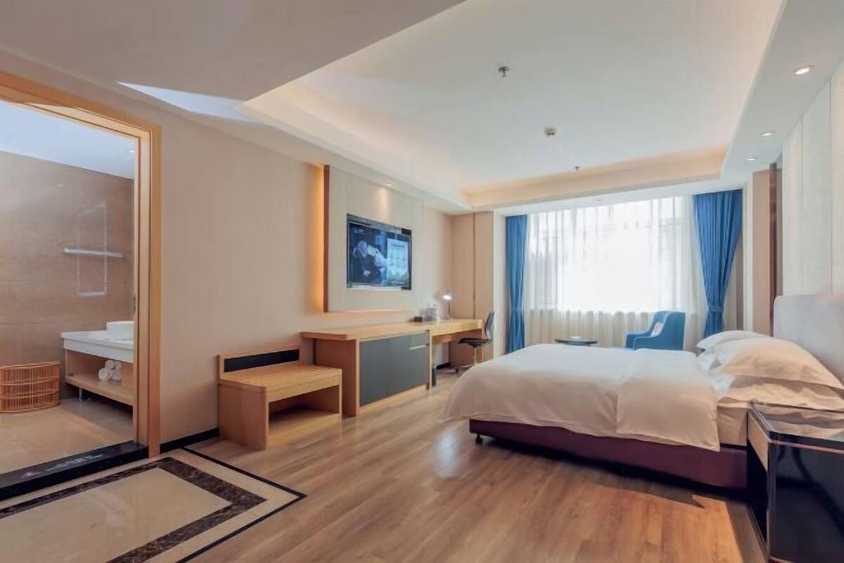 A modern hotel room with a large bed, a desk, a chair, a TV, and a bathroom with a shower and toilet.
