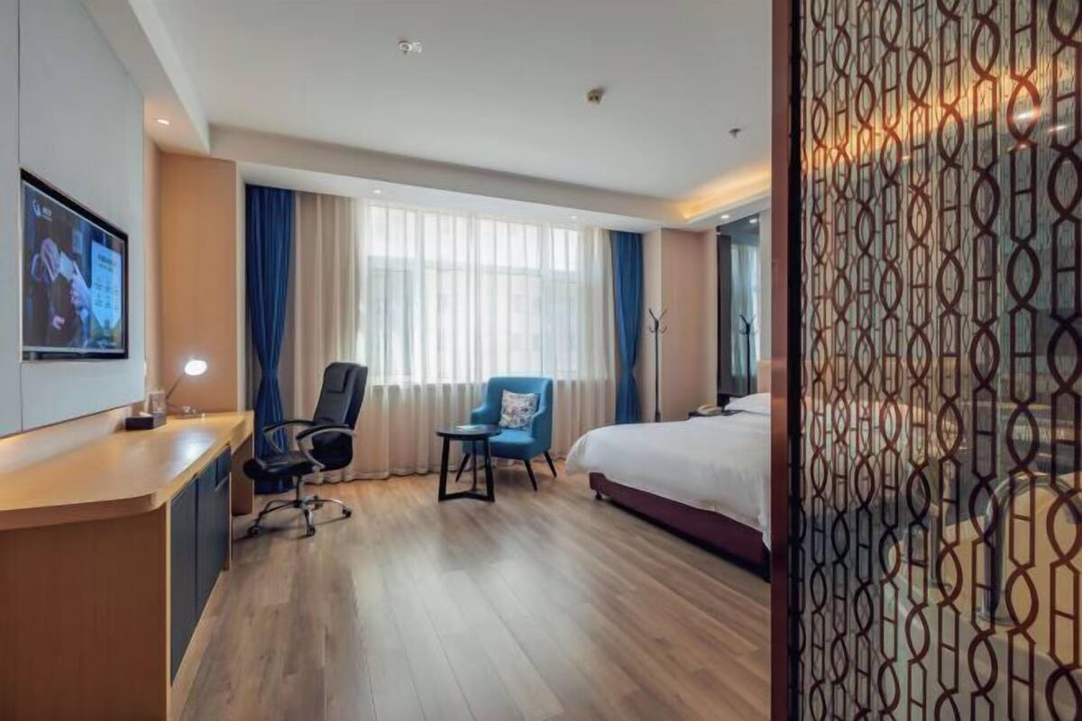 A hotel room with a wooden floor, a desk with a lamp, a bed, a chair, and a TV on the wall.