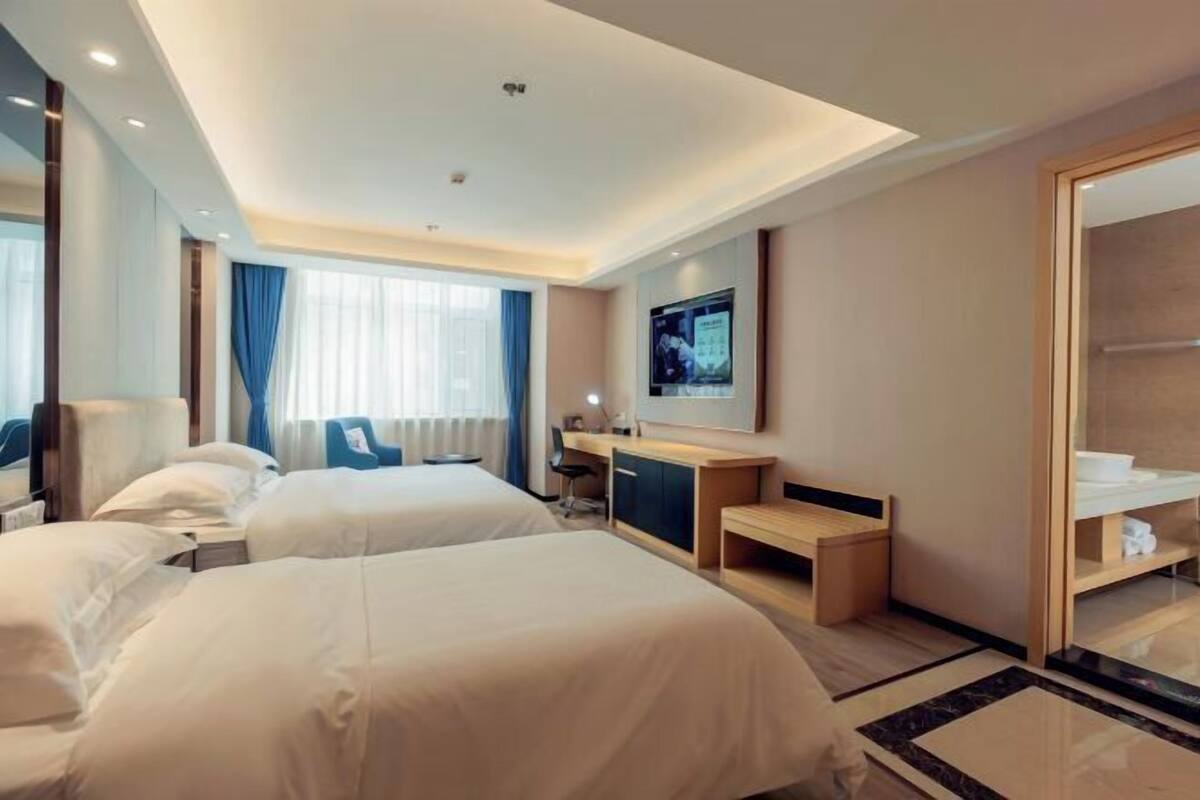 A hotel room with two beds, a desk, a chair, a TV, and a bathroom with a mirror and towel rack.