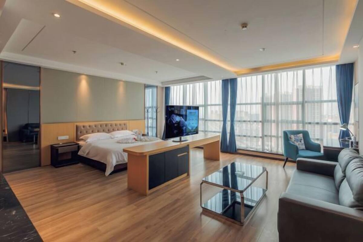 A modern hotel room with a bed, a TV, a sofa, a glass coffee table, and large windows.