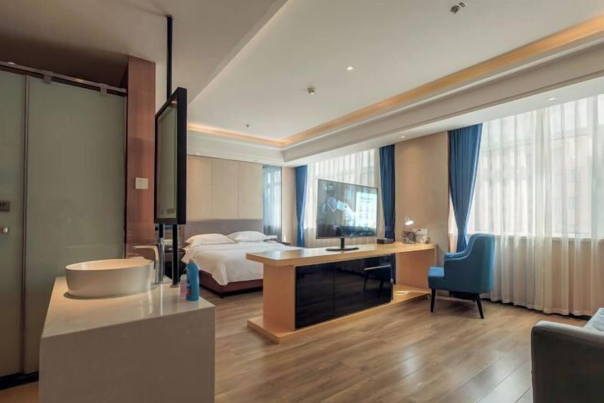 A modern hotel room with a bed, a TV, a desk, a chair, and a large window with curtains.