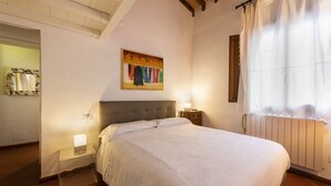 A bedroom with a bed, bedside lamps, a window with curtains, a radiator, and a colorful painting on the wall.