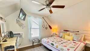 A bedroom with a bed, a desk, a chair, a ceiling fan, a window with curtains, and a wall-mounted lamp.