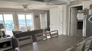 A dining area with a large wooden table and white chairs, a ceiling fan, and a view of the ocean through large windows. - Beach Front with Charm and Luxury. 2 person Hot Tub on Deck. Beautiful Loft! (North Topsail Beach)