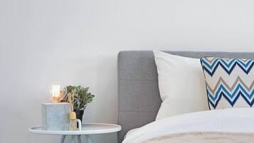A neatly arranged bedroom with a grey headboard, a white pillow with a zigzag pattern, a bedside table with a lamp, and a small potted plant.