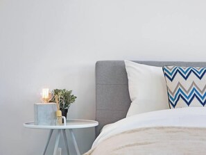 Une chambre à coucher bien rangée, avec une tête de lit grise, un oreiller blanc à motifs en zigzag, une table de chevet avec une lampe et une petite plante en pot.