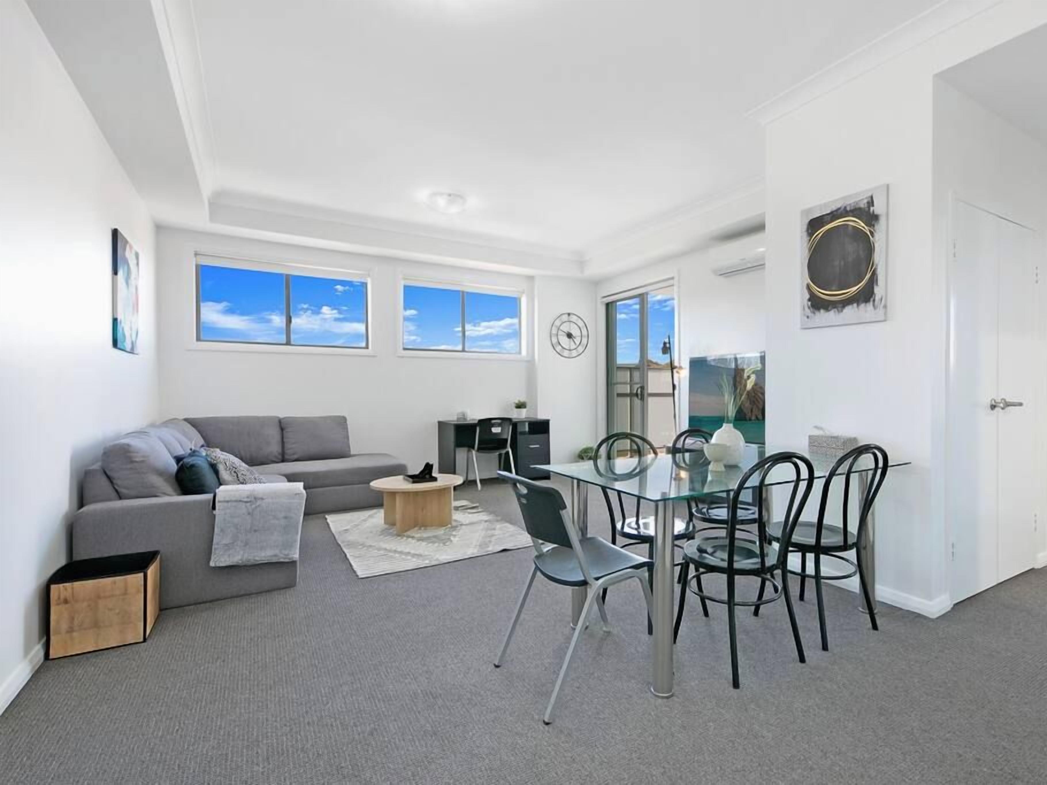 A modern living room with a grey sofa, a glass dining table with chairs, and a small wooden side table. There are windows with a view of the sky, a door, and a wall-mounted artwork.