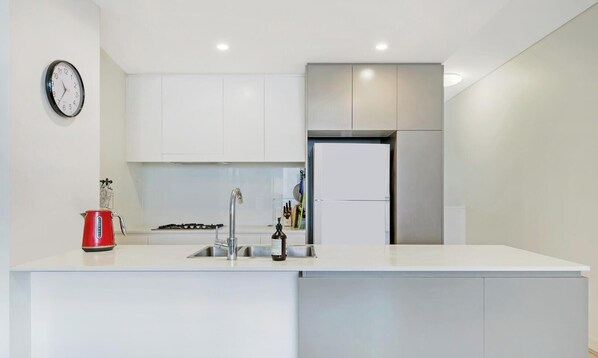 Una cocina moderna con muebles blancos, un refrigerador de acero inoxidable, un hervidor rojo y un reloj de pared.