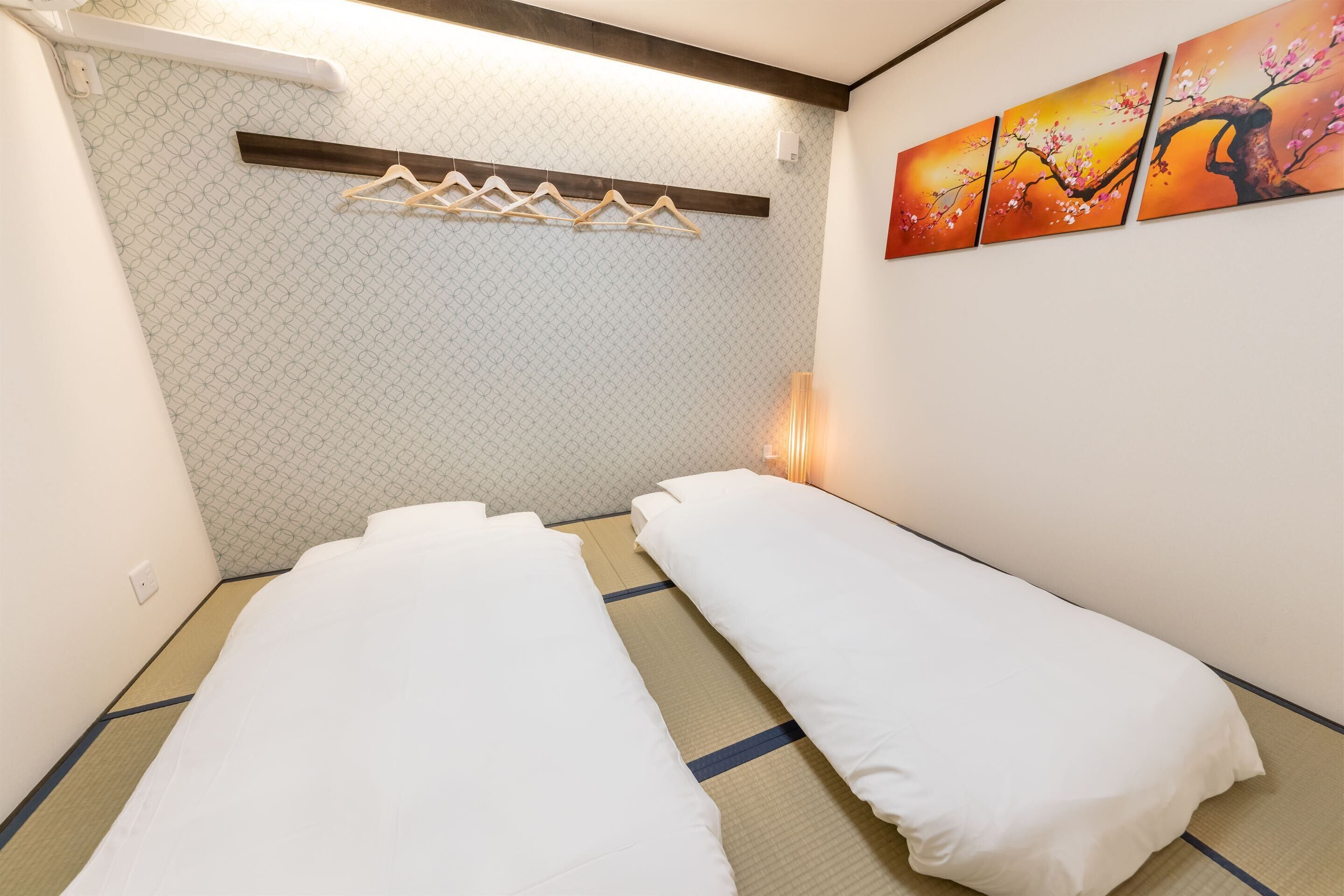 A room with two single beds, a wooden shelf with hangers, a wall-mounted light, and a painting of cherry blossoms.
