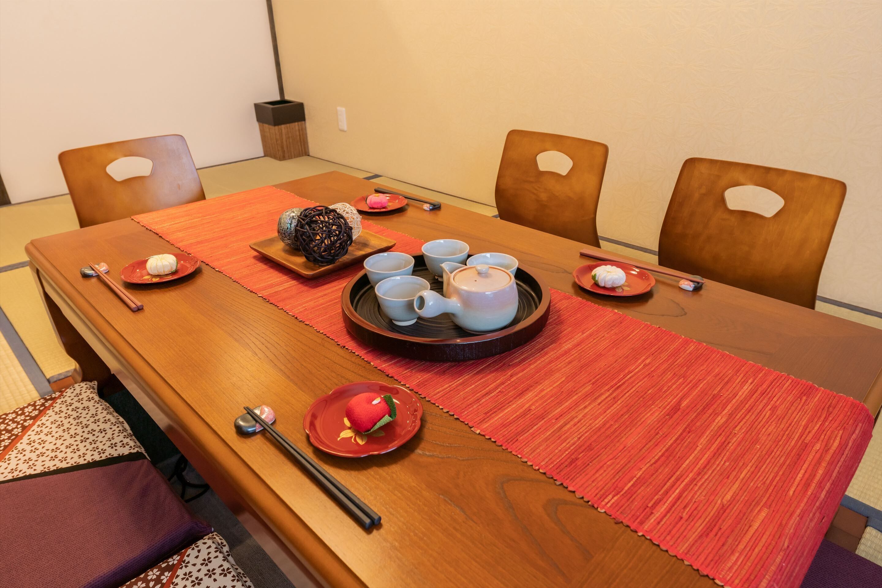 Una mesa de comedor de madera con un mantel individual rojo, rodeada por cuatro sillas. Hay platos con comida y una tetera sobre la mesa.