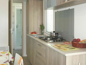 A modern kitchen with a gas stove, sink, and wooden cabinets. There is a window with blinds, a potted plant, and a basket of fruit on the counter.