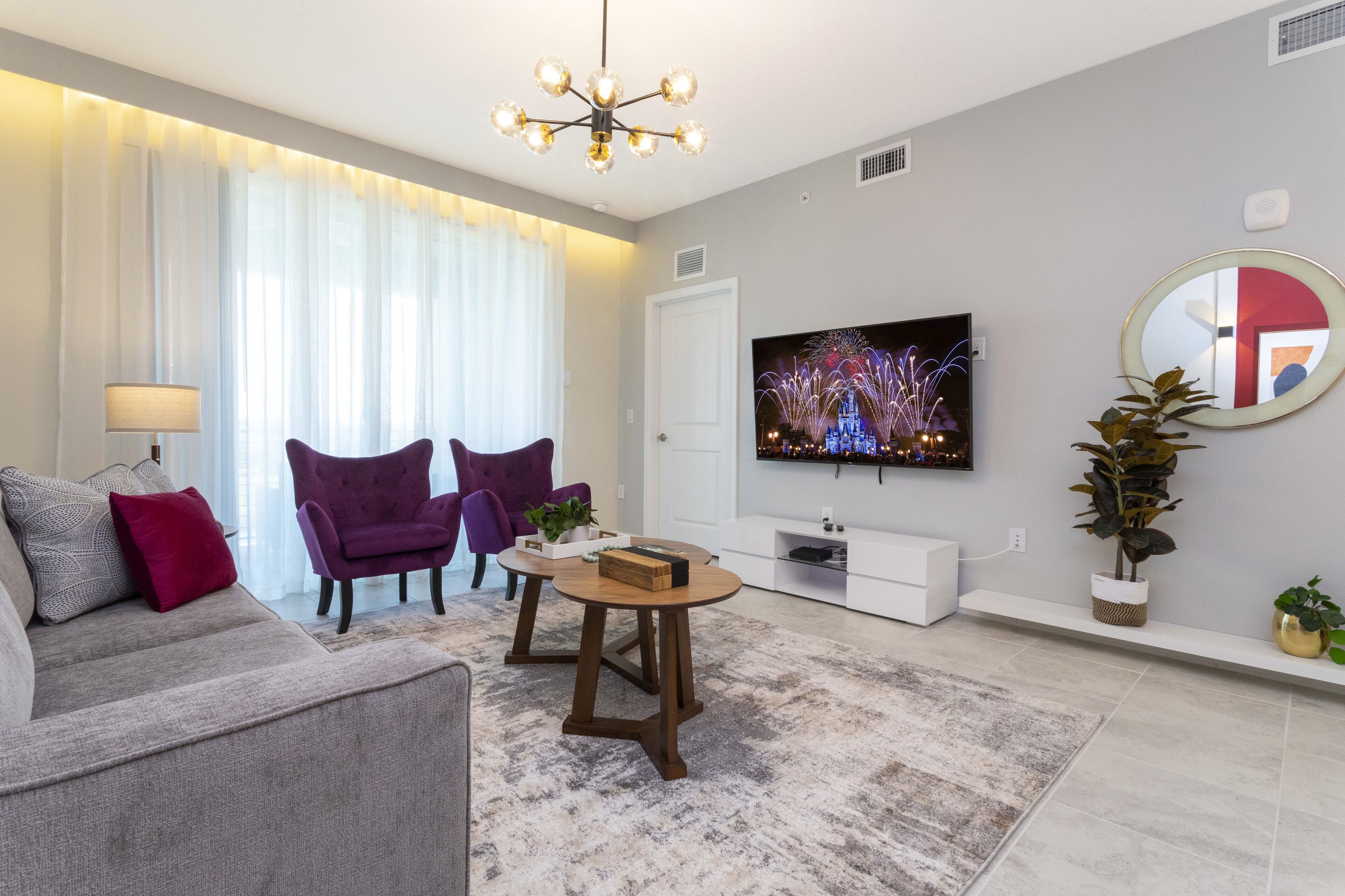 A modern living room with a grey sofa, two purple armchairs, a wooden coffee table, a flat-screen TV, and a large mirror.