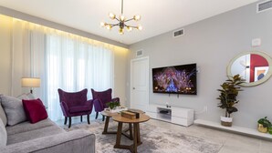 Ein modernes Wohnzimmer mit einem grauen Sofa, zwei violetten Sesseln, einem Holztisch, einem Flachbildfernseher und einem großen Spiegel.