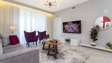 A modern living room with a grey sofa, two purple armchairs, a wooden coffee table, a flat-screen TV, and a large mirror.