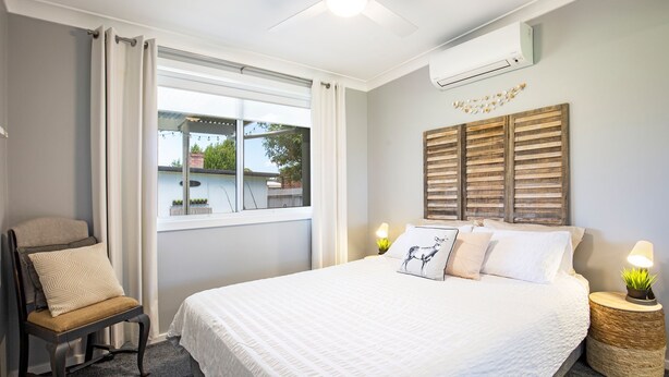 Ein Schlafzimmer mit einem Bett, einem Sessel, einem Fenster mit Vorhängen, einer Wandklimaanlage und einem Wandbild.