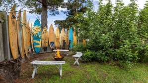 Ein Holzzaun mit Surfbrettern, einer Feuerstelle und zwei weißen Stühlen in einer grünen Außenanlage.