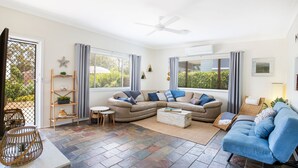 Ein geräumiges Wohnzimmer mit einer großen, L-förmigen Couch, einem blauen Sessel und einem kleinen runden Tisch. Zwei Fenster mit Jalousien, ein Deckenventilator und eine Holzleiter mit Pflanzen sind ebenfalls vorhanden.