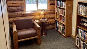 A cozy log cabin room with a large window, a wooden chair, a small table with a lamp, and two bookshelves filled with books. - Rustic log cabin (Naples)