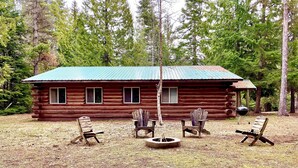 A log cabin with a metal roof, surrounded by a grassy area with four wooden chairs and a fire pit. - Rustic log cabin (Naples)