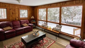 Interior acogedor de una cabaña con sofá de cuero, una mesita de centro y grandes ventanales que dan a una vista nevada de un bosque.