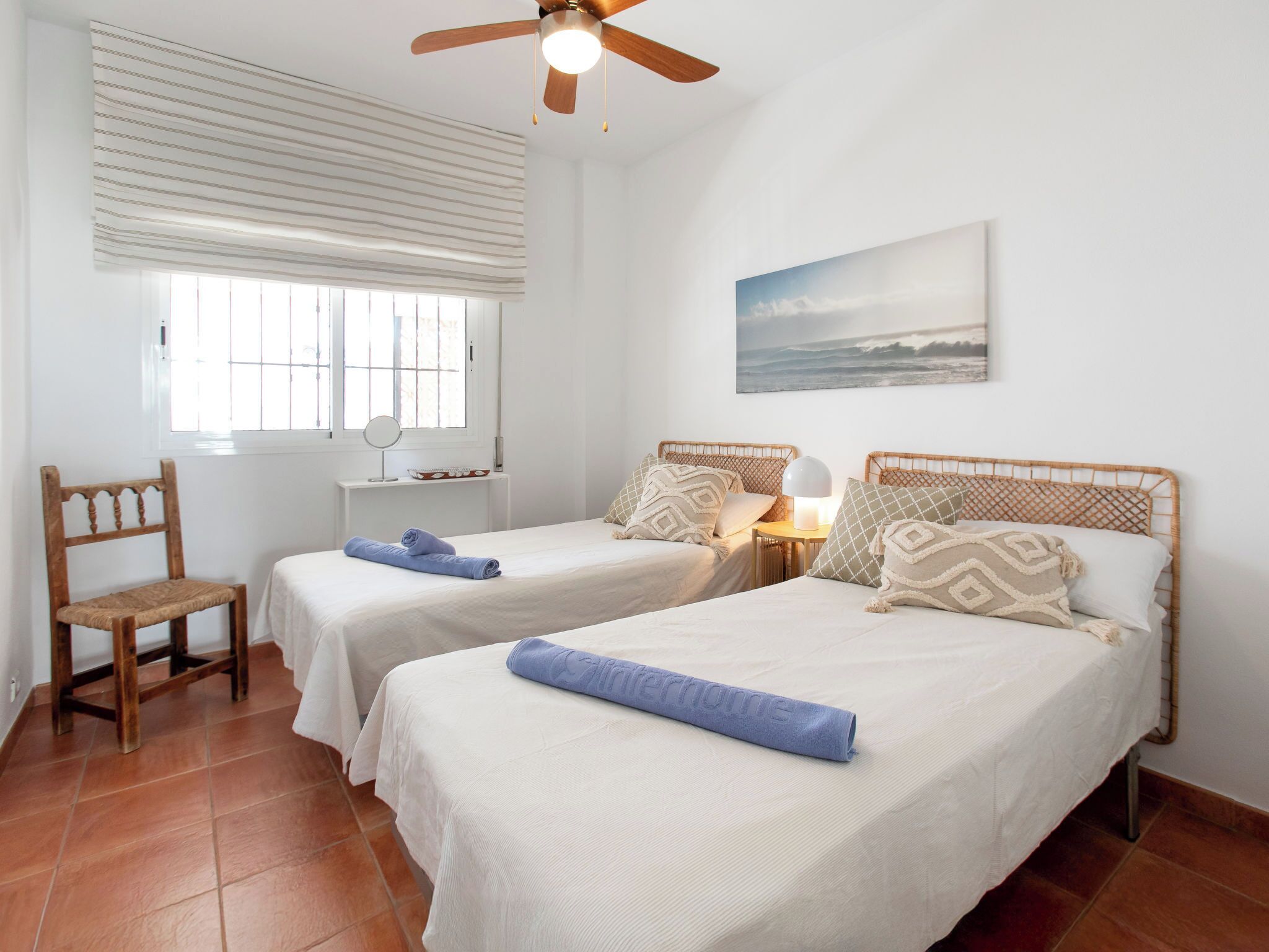 A room with two beds, a chair, a desk, a ceiling fan, and a window with blinds.
