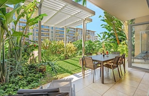 A patio with a table and chairs, surrounded by lush greenery and a view of a multi-story building.