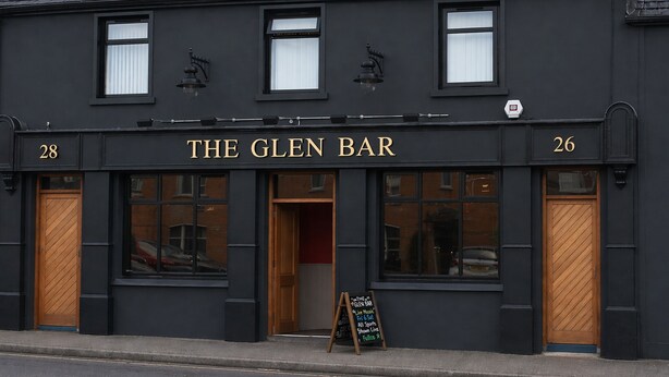 En svart byggnad med skylten ”THE GLEN BAR”, två fönster och en trädörr.
