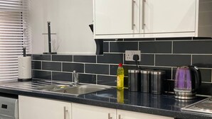 A kitchen with white cabinets, a black countertop, a stainless steel kettle, and a coffee maker. - 2 Bed Flat - Sleeps 6 - Parking - Wifi (Bridgend)