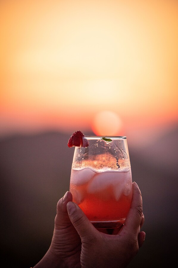A person holding a glass with a red drink and a strawberry garnish against a sunset background. - Starview Ranch: 3 homes w/ 7 bdrms, pool, sauna, spa & gym on 50 beautiful acres (Miramonte)