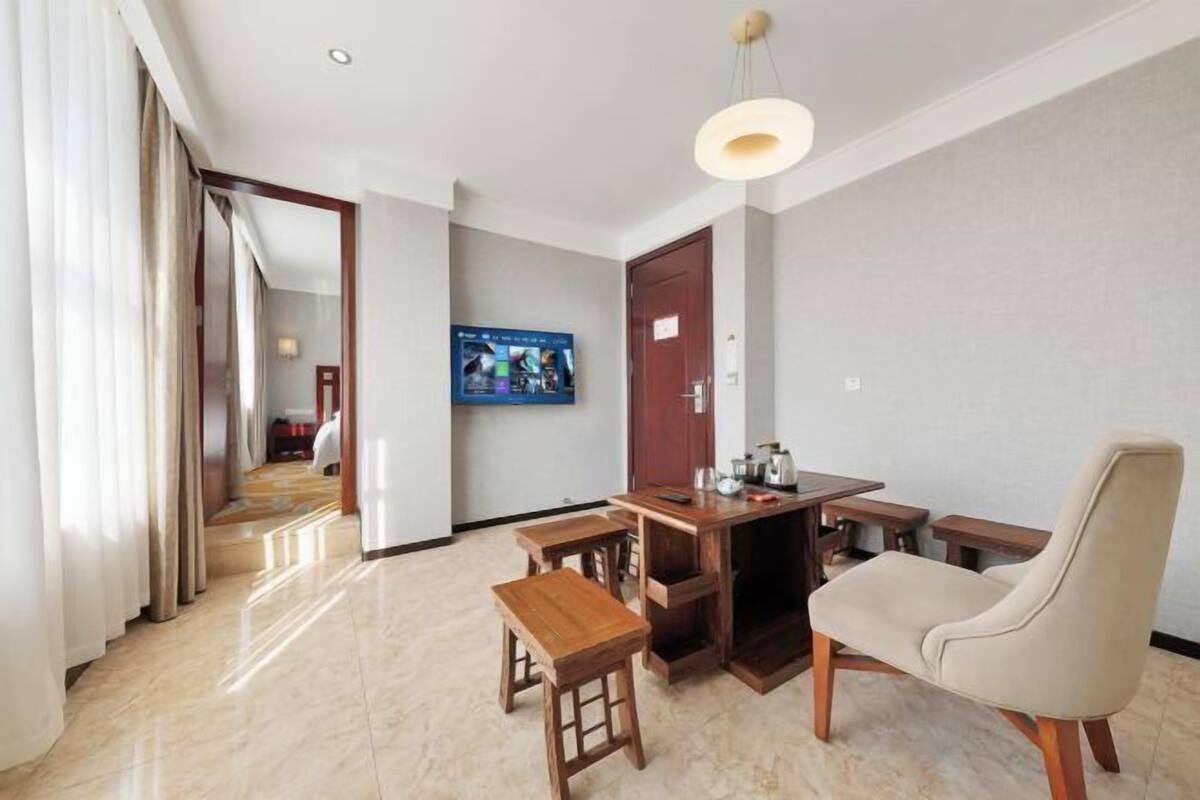 A hotel room with a wooden dining table, two wooden stools, a beige armchair, and a television mounted on the wall.