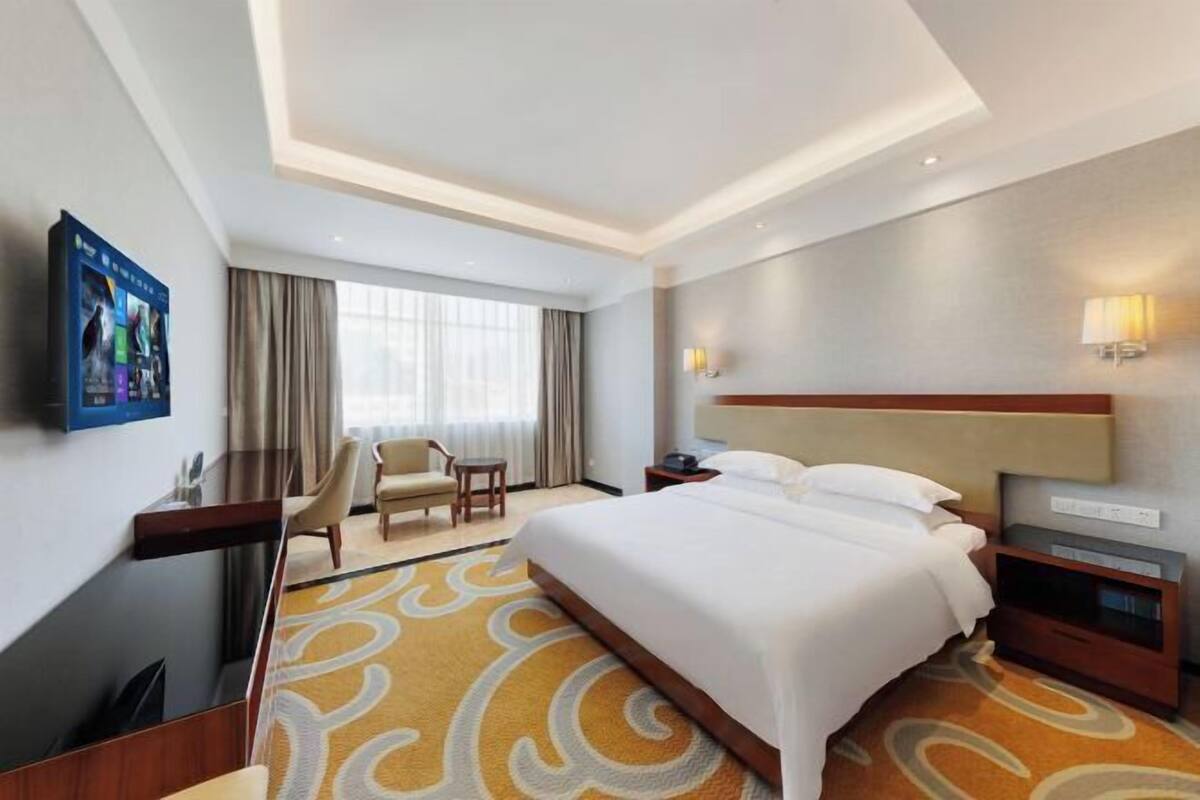 A hotel room with a large bed, a desk with a chair, a TV mounted on the wall, and a window with curtains.