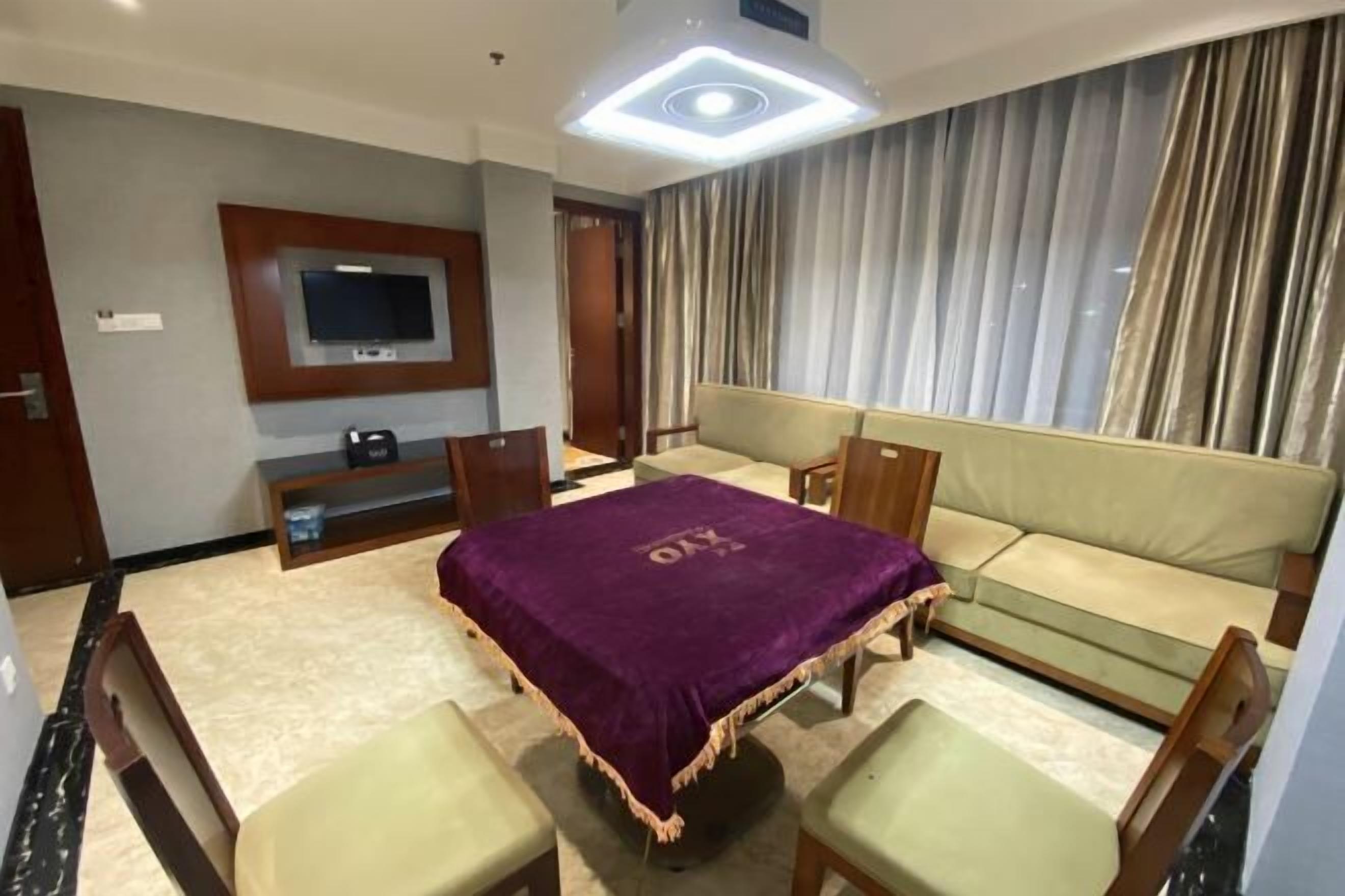 A hotel room with a sofa, a table with a purple tablecloth, a TV mounted on the wall, and a window with curtains.