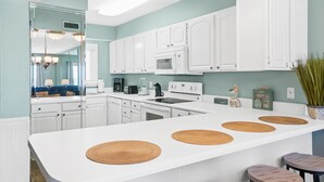 A modern kitchen with white cabinets, a central island with woven placemats, and a dining area with stools. - 2nd Floor Beachfront Getaway | Regency Towers, Pools, Tiki Bar & Gulf Views (Panama City)