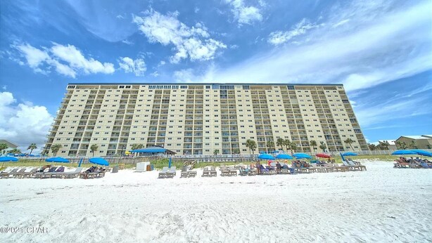 A large hotel with multiple floors and balconies, situated on a sandy beach with beach umbrellas and lounge chairs. - 2nd Floor Beachfront Getaway | Regency Towers, Pools, Tiki Bar & Gulf Views (Panama City)